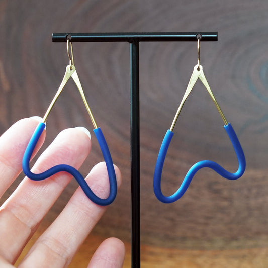 A pair of blue MCM style earrings with brass wire and gold-filled hooks, displayed on a black T-stand with a hand holding one earring for scale against a dark wood background by artist Jacki Geary with JAX Atelier.