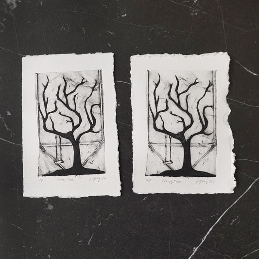 Black and white tetra pak drypoint intaglio limited edition print featuring a stylized creepy tree with a swing, and torn white deckled paper edges, displayed side by side on a dark marble surface. By Jacki Geary with Jax Atelier.