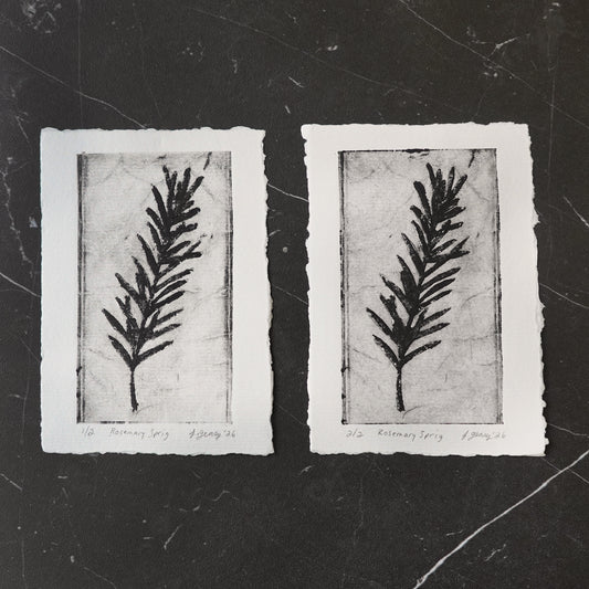 Two black and white botanical tetra pak drypoint intaglio prints of a rosemary sprig on thick watercolor paper with a deckled edge, displayed on a dark textured slate background. By artist Jacki Geary with Jax Atelier in San Diego.