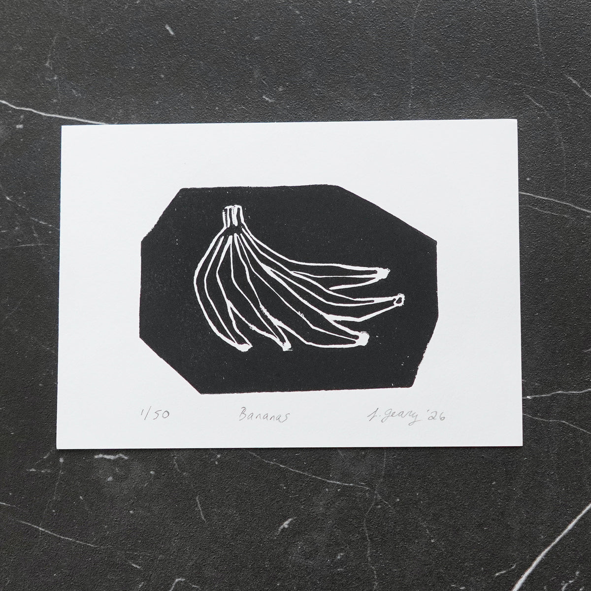 A black and white linocut art print depicting a bunch of bananas, displayed on a dark textured marble surface. By Jax Atelier in San Diego.