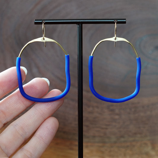 A pair of large square abstract hoop earrings with bright blue rubber lower sections and brass upper area, hanging from a black T-stand against a blurred brown wood-grain background and a hand is holding one earring on the left for scale against a blurred brown wood background, made by Jacki Geary with Jax Atelier.
