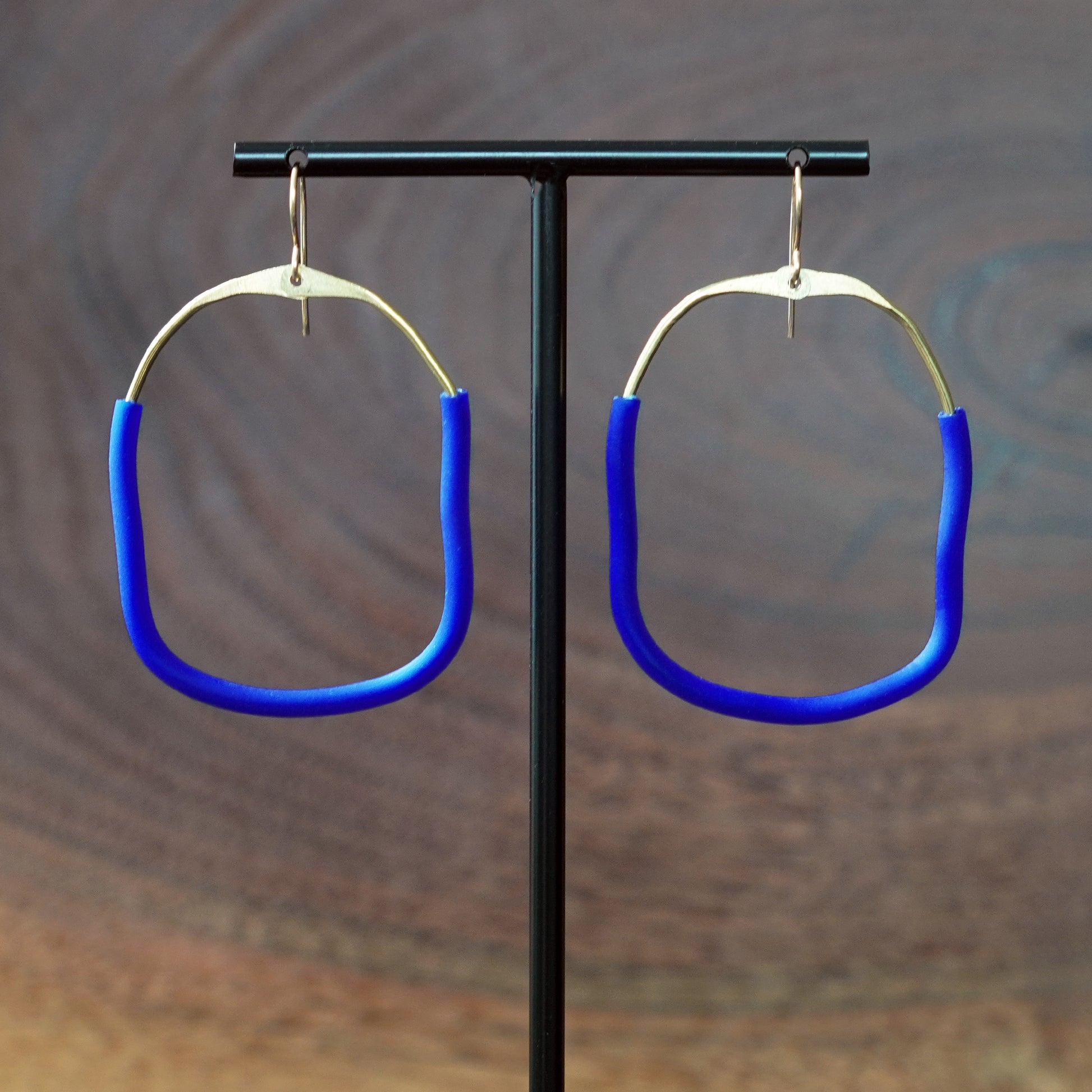 A pair of large square abstract hoop earrings with bright blue rubber lower sections and brass upper area, hanging from a black T-stand against a blurred brown wood-grain background, made by Jacki Geary with Jax Atelier.