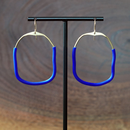 A pair of large square abstract hoop earrings with bright blue rubber lower sections and brass upper area, hanging from a black T-stand against a blurred brown wood-grain background, made by Jacki Geary with Jax Atelier.
