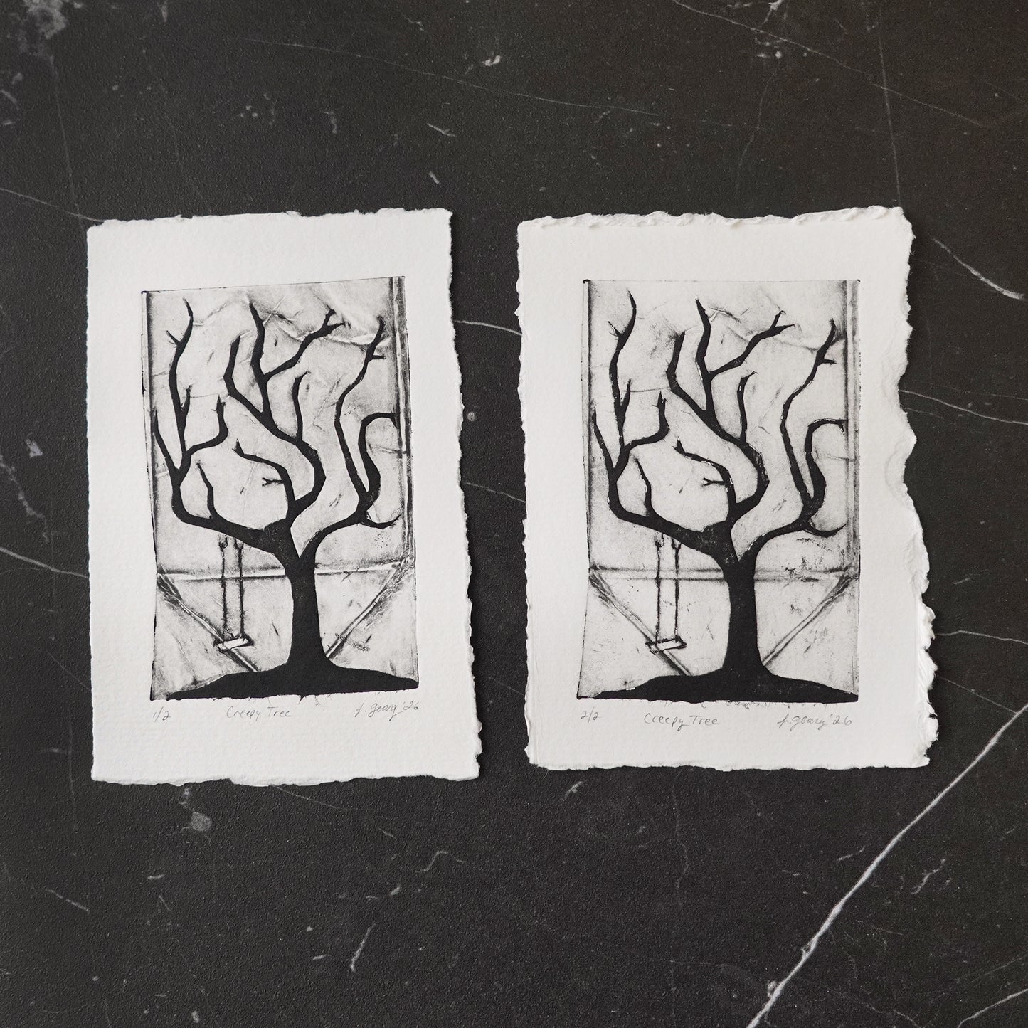 Black and white tetra pak drypoint intaglio limited edition print featuring a stylized creepy tree with a swing, and torn white deckled paper edges, displayed side by side on a dark marble surface. By Jacki Geary with Jax Atelier.