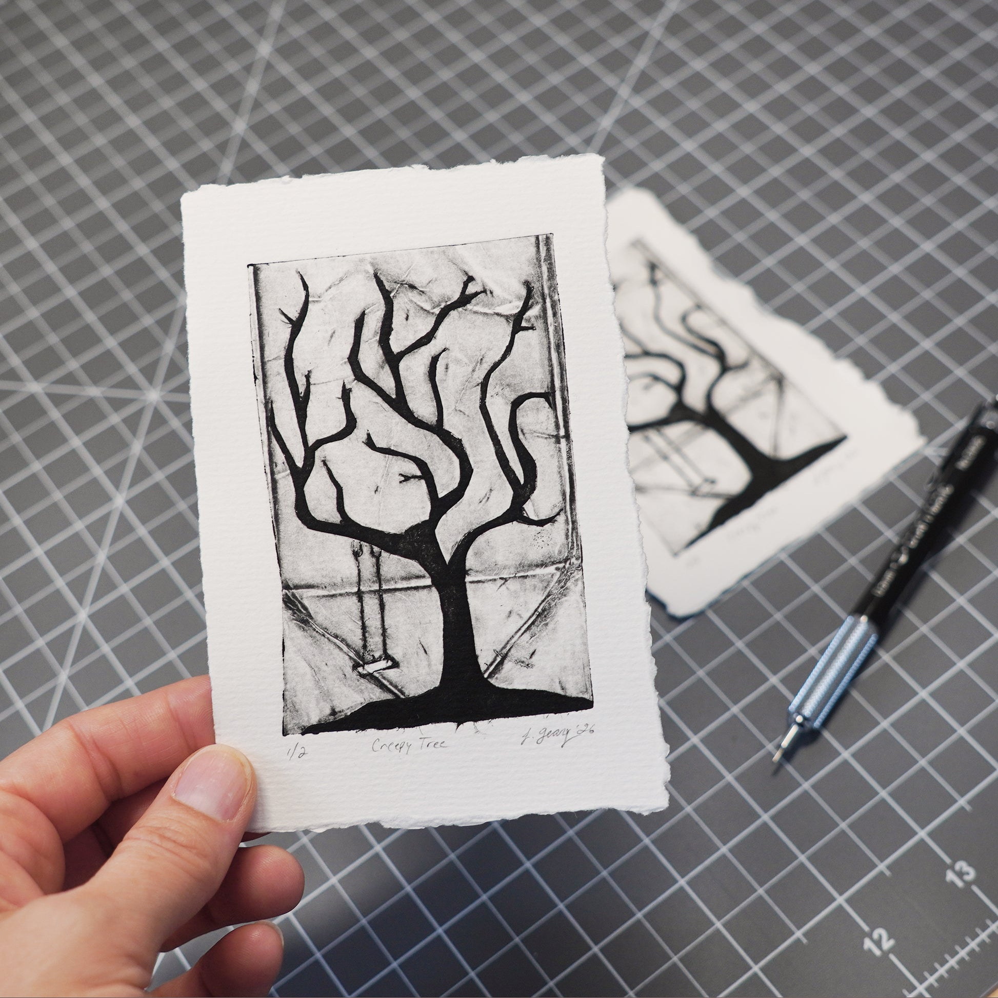 A hand holds a small black-and-white tetra pak drypoint print featuring a tree with no leaves and a hanging swing, shown over a gray gridded cutting mat with a second print and a pencil in the background. Art by Jacki Geary with JAX Atelier.