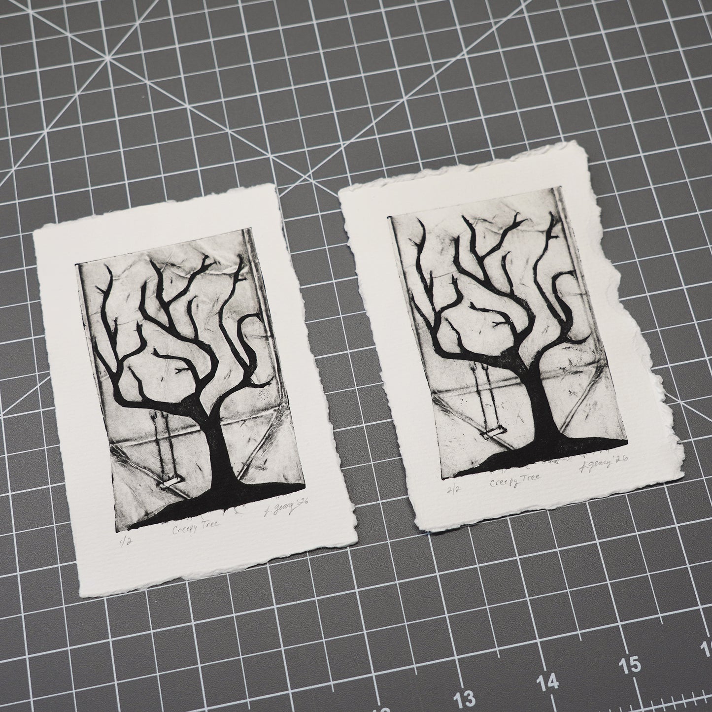 Two handmade tetra pak drypoint prints on rough white deckled edge paper, featuring a black ink etching of a tree with hanging swing, displayed on a gray cutting mat with grid lines. Art by Jax Atelier.