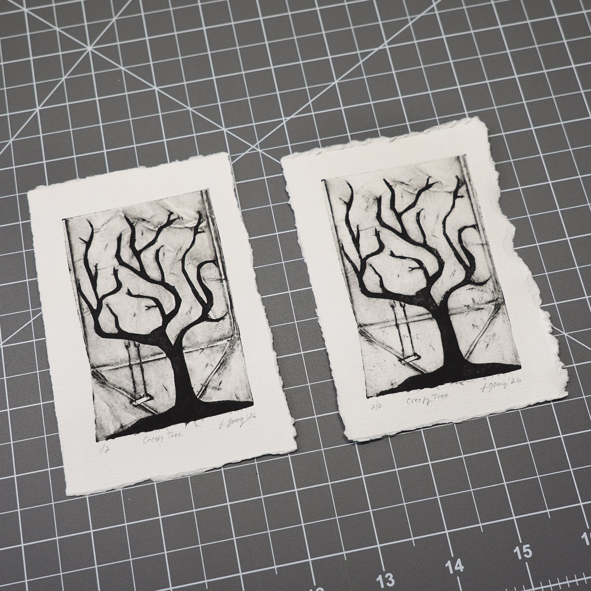Two handmade tetra pak drypoint prints on rough white deckled edge paper, featuring a black ink etching of a tree with hanging swing, displayed on a gray cutting mat with grid lines. Art by Jax Atelier.