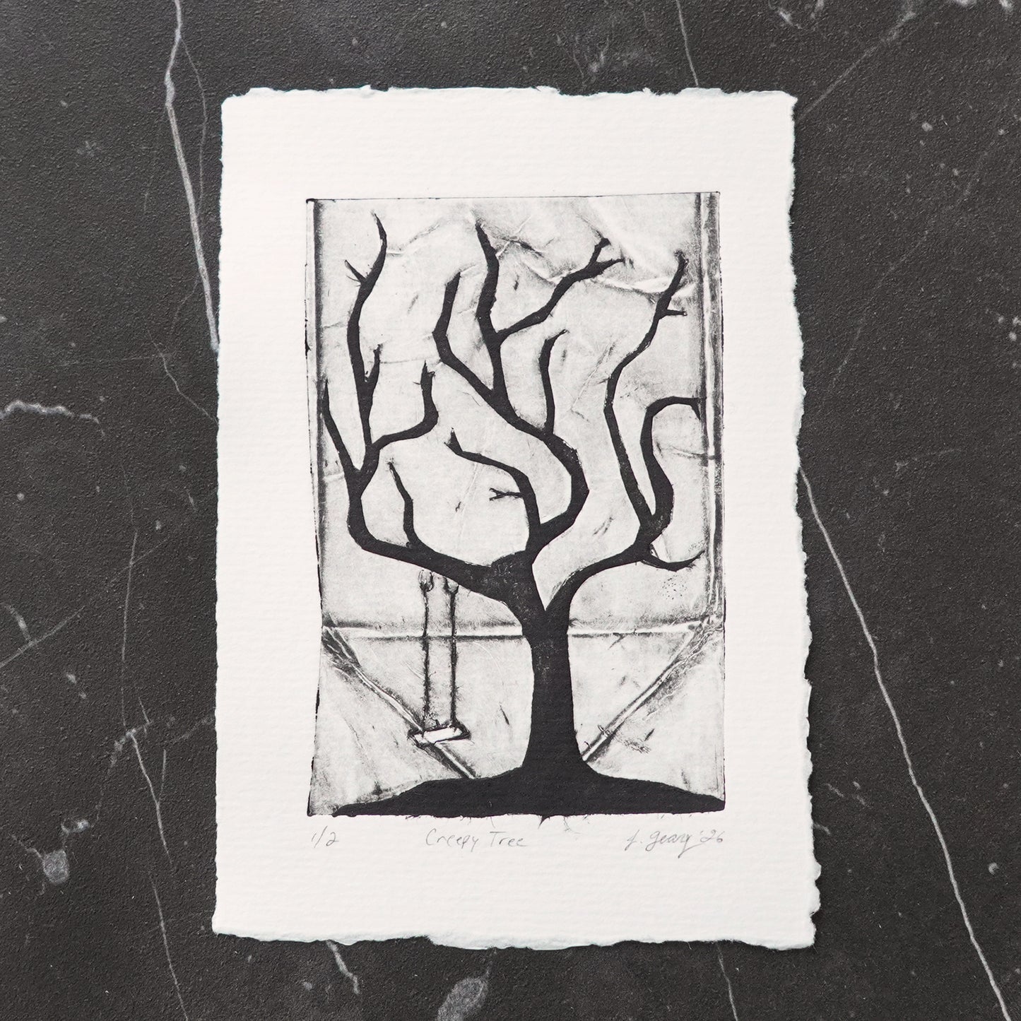 A black and white tetra pak drypoint intaglio limited edition print featuring a stylized creepy tree with a swing, displayed on a dark marble surface. By Jacki Geary with Jax Atelier.
