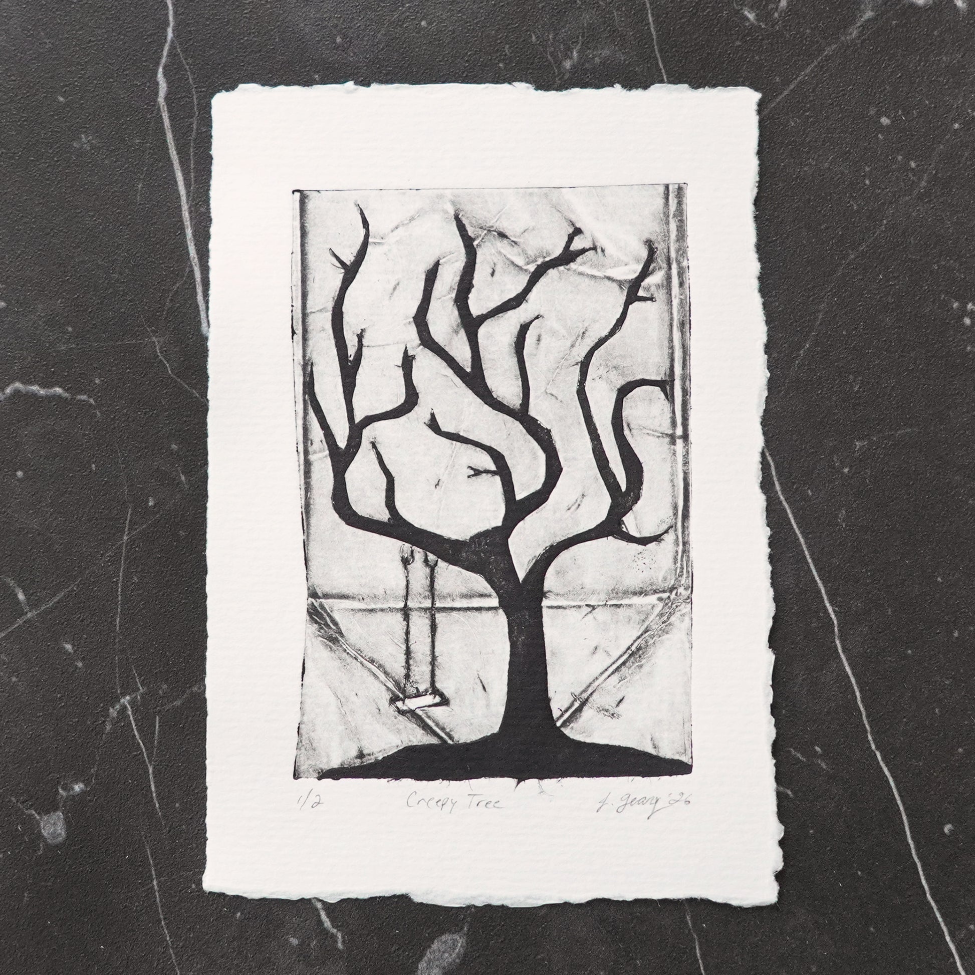 A black and white tetra pak drypoint intaglio limited edition print featuring a stylized creepy tree with a swing, displayed on a dark marble surface. By Jacki Geary with Jax Atelier.