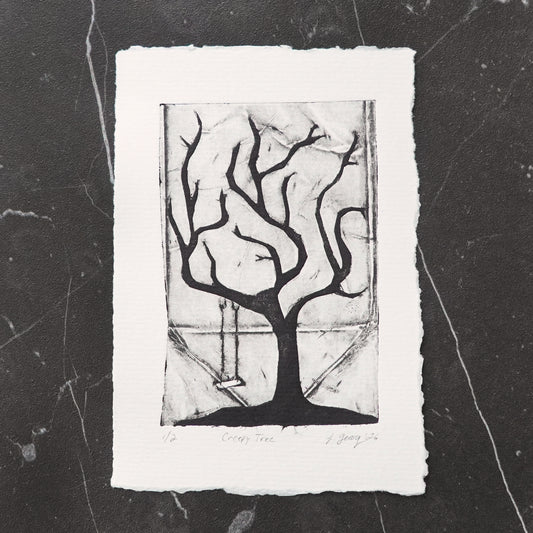 A black and white tetra pak drypoint intaglio limited edition print featuring a stylized creepy tree with a swing, displayed on a dark marble surface. By Jacki Geary with Jax Atelier.