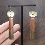 A pair of handmade dangle geometric earrings displayed on a black T-stand against a dark wood grain background. The earrings feature a hammered gold-tone brass top disc riveted to a turquoise blue patina piece. A vertical patinated copper bar hangs from the top piece of the earring. Ear hooks are made with 14k gold-filled wire. Earrings are handmade by JAX Atelier in San Diego, CA.