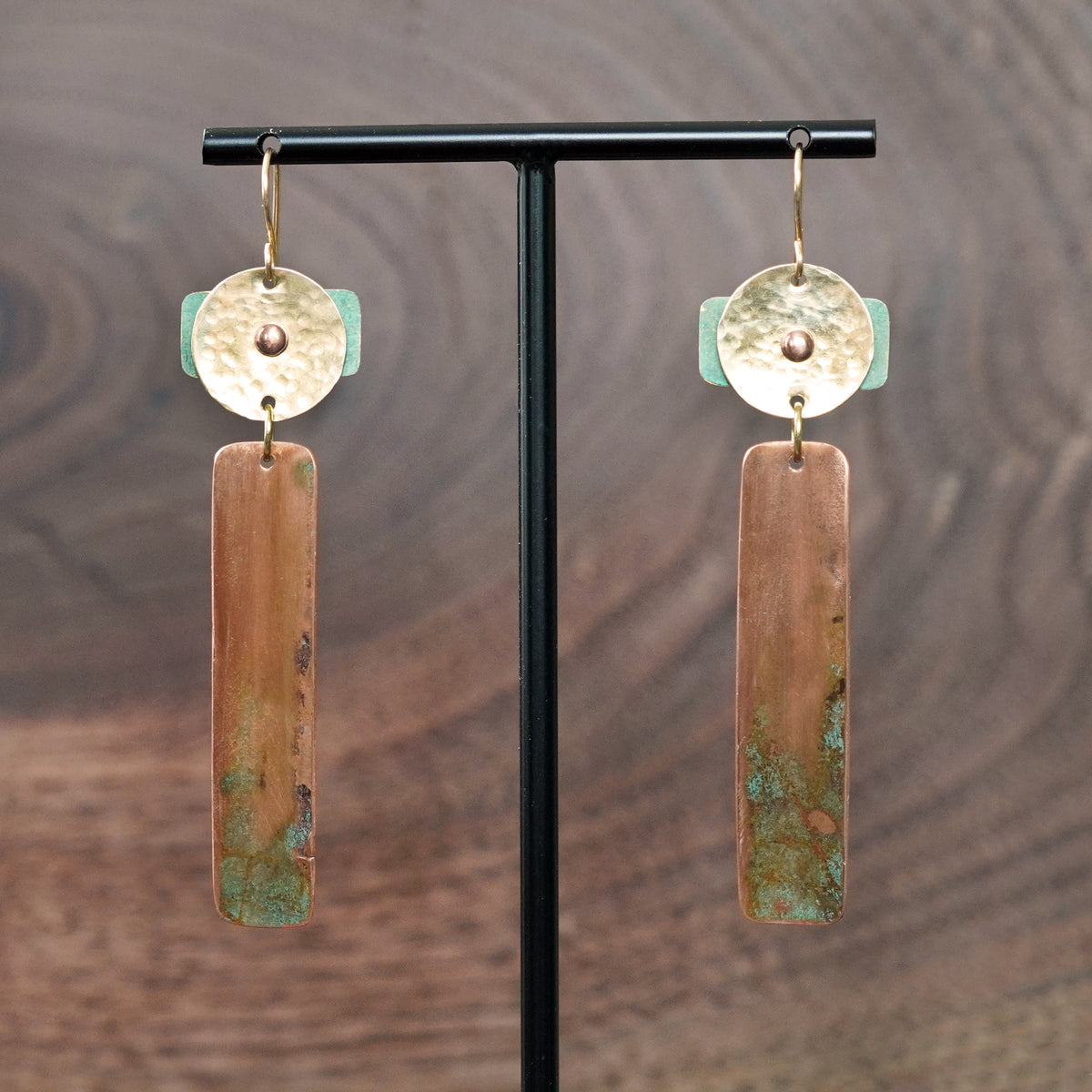 A pair of handmade dangle geometric earrings displayed on a black T-stand against a dark wood grain background. The earrings feature a hammered gold-tone brass top disc riveted to a turquoise blue patina piece. A vertical patinated copper bar hangs from the top piece of the earring. Ear hooks are made with 14k gold-filled wire. Earrings are made by JAX Atelier in San Diego, CA.