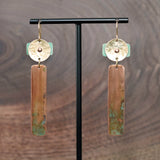 A pair of handmade dangle geometric earrings displayed on a black T-stand against a dark wood grain background. The earrings feature a hammered gold-tone brass top disc riveted to a turquoise blue patina piece. A vertical patinated copper bar hangs from the top piece of the earring. Ear hooks are made with 14k gold-filled wire. Earrings are made by JAX Atelier in San Diego, CA.