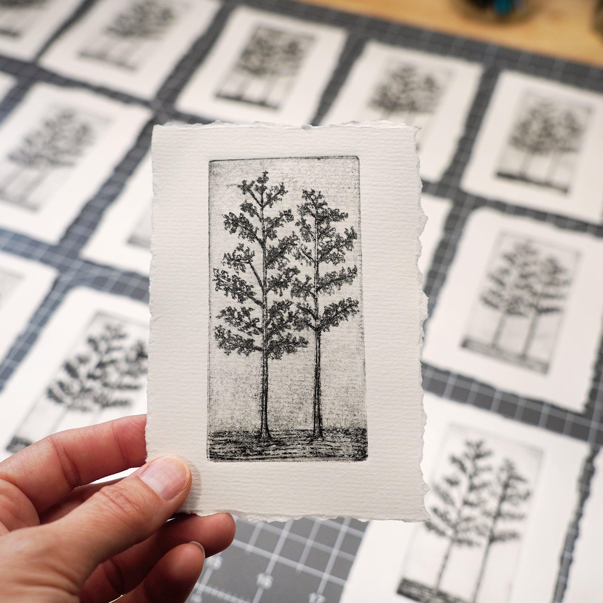 A hand holds a small art print featuring a black-and-white etched tree design with torn paper edges. Multiple similar prints are laid out on a cutting mat in the blurred background. By Jax Atelier in San Diego.