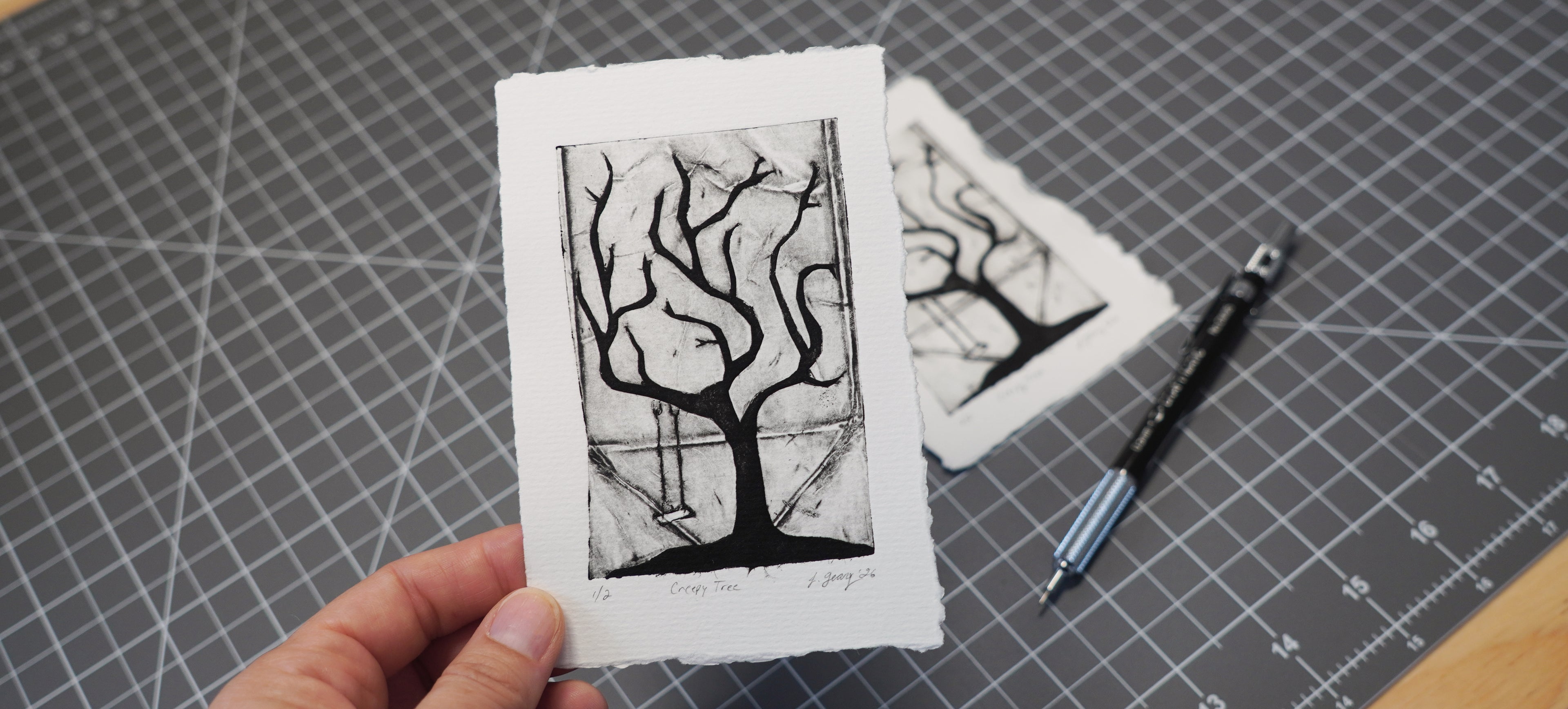 A hand holds a black and white Tetra Pak art print featuring a creepy tree with a swing, set against a dark gray cutting mat with a second print and a pencil in the background. By Jax Atelier in San Diego.