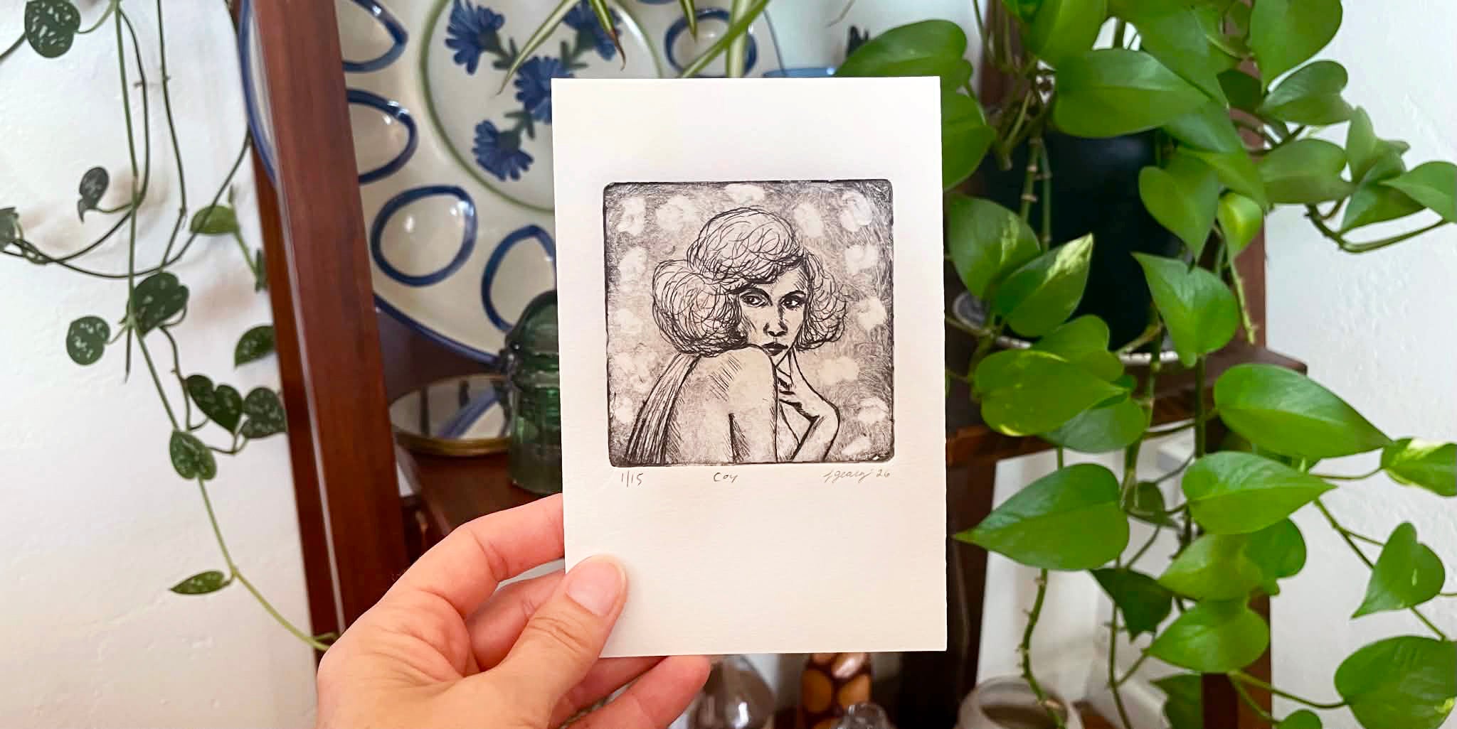 A hand holds a white drypoint intaglio print featuring a black ink sketch of a woman, with green houseplants and a decorative plate in the background. By Jax Atelier in San Diego.
