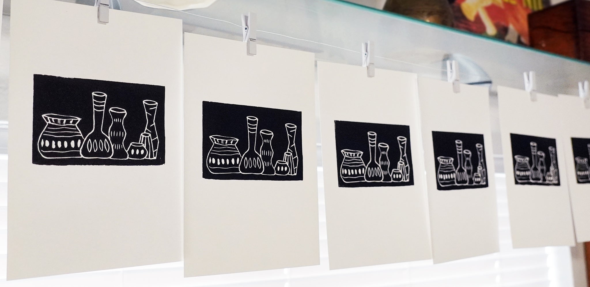 A row of white linocut prints clipped along a glass shelf, each featuring a black square panel with a white line drawing of decorative vases and jars. By Jax Atelier in San Diego.