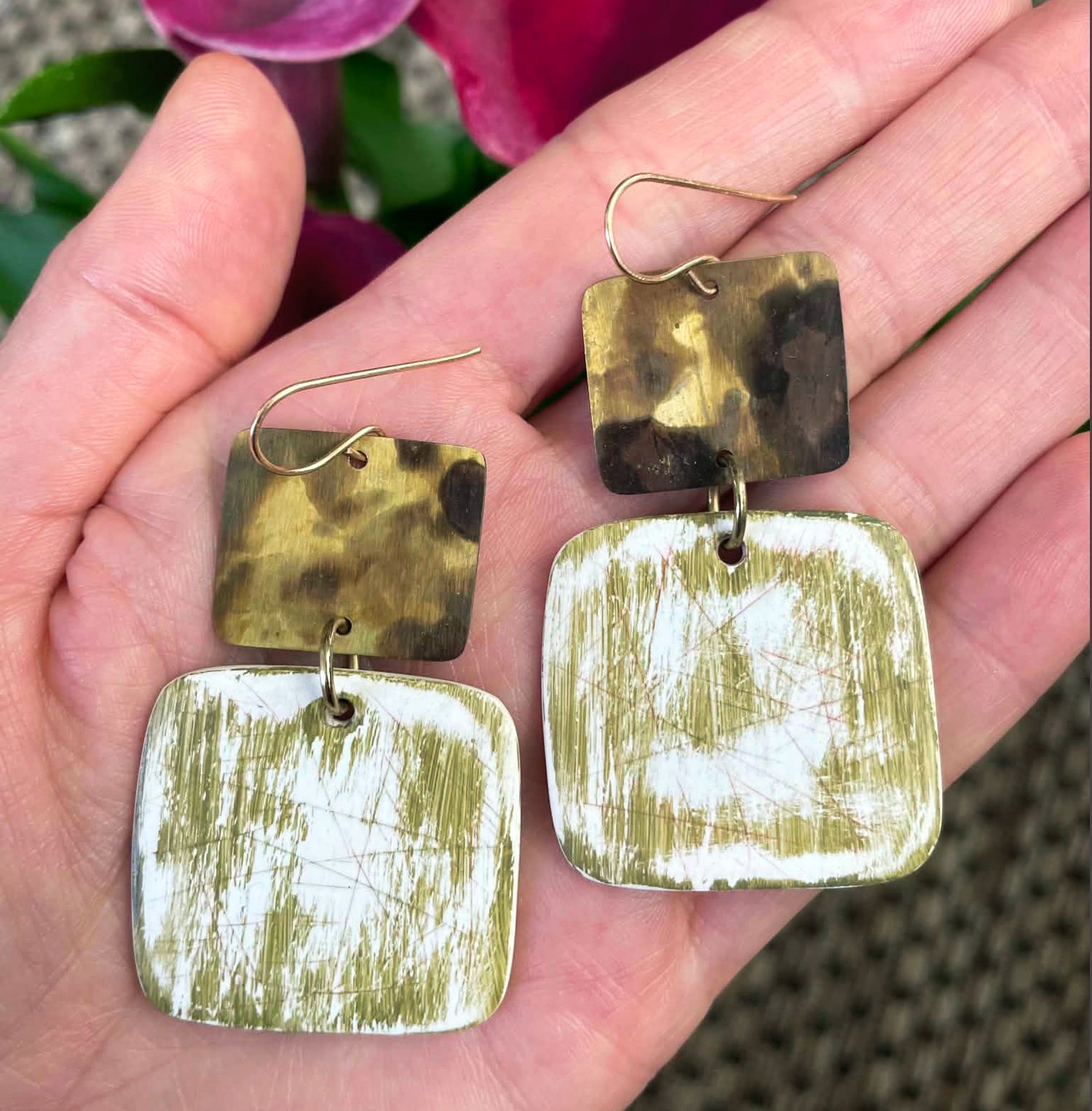 A pair of handmade square drop earrings held in a hand. Each earring has a small gold-toned patina square with a dark marbled section, connected to a larger distressed white and olive green square. By Jax Atelier in San Diego.