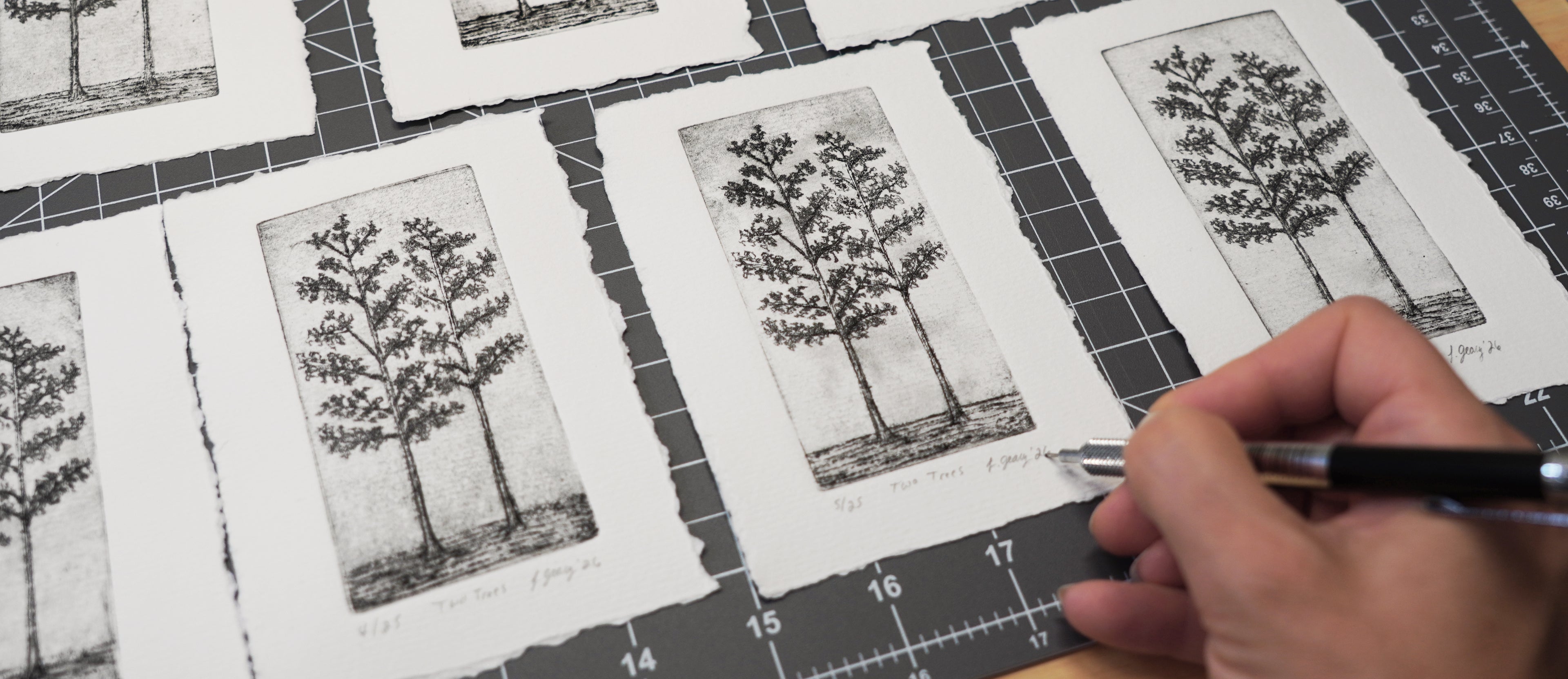A close-up of a hand holding a pen over several printed tree drypoint etchings laid out on a dark gridded cutting mat. By Jax Atelier in San Diego.