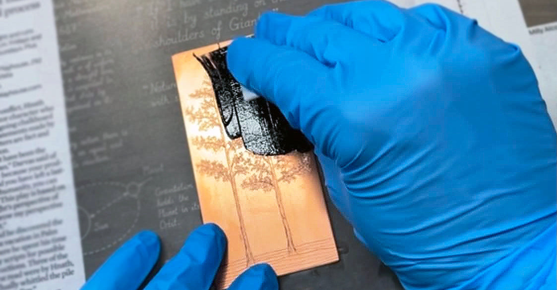 A close-up of a person wearing blue nitrile gloves applying black ink to a copper plate with engraved tree designs, set against a dark background with printed papers. By Jax Atelier in San DIego.