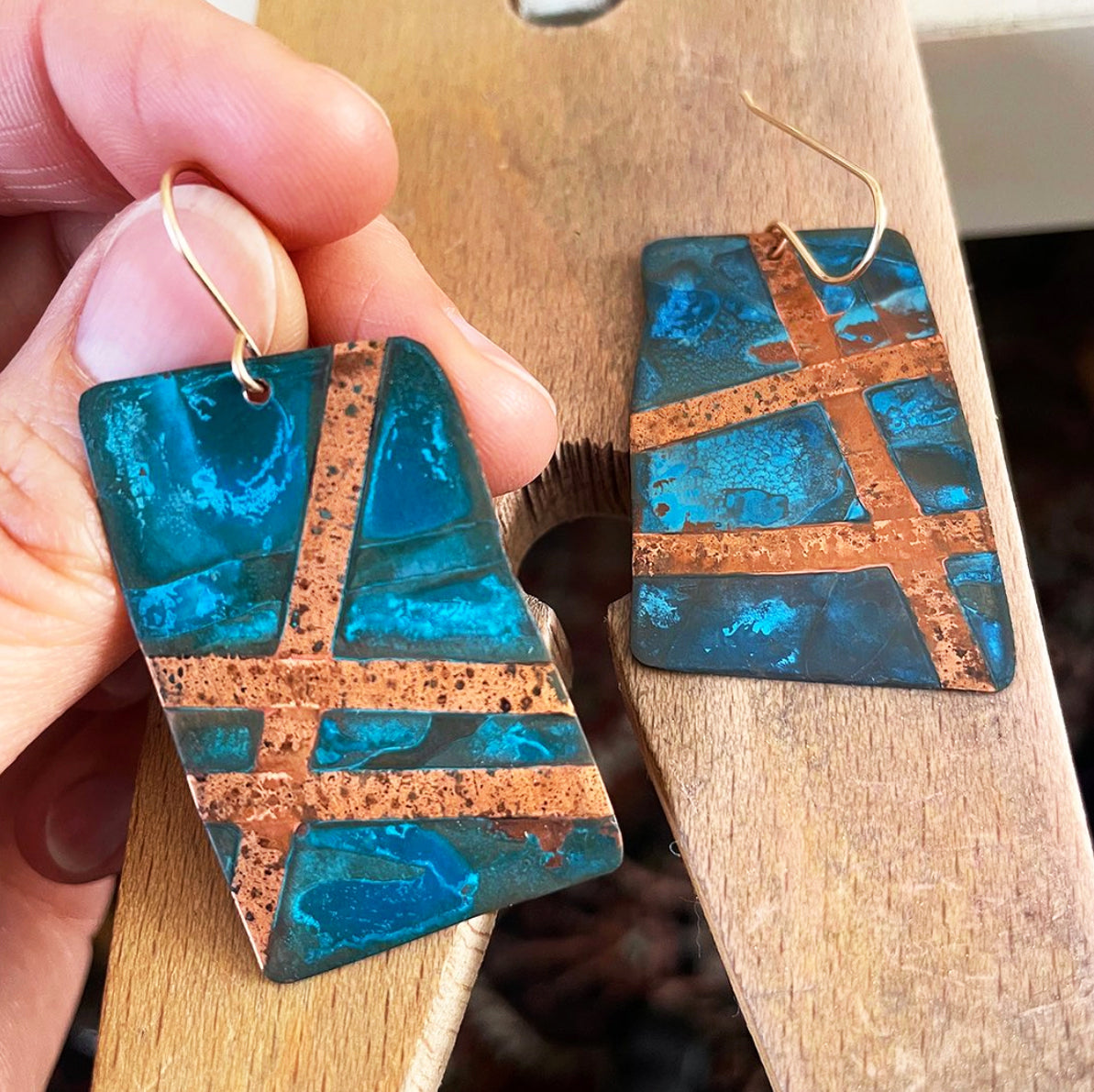 A close-up of a hand holding a pair of rectangular drop earrings with a textured turquoise blue patina finish and copper cross patterns, with the second earring resting on a wooden surface. Made by JAX Atelier in San Diego, CA.