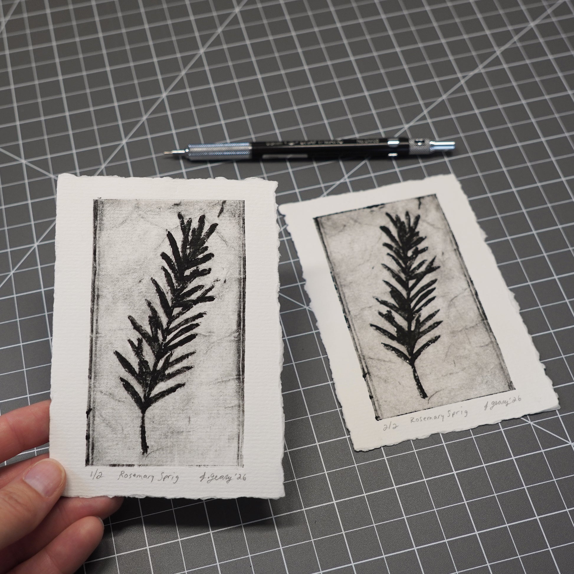 Two handmade botanical limited edition tetra pak drypoint prints with black ink rosemary illustrations are shown on a gray gridded cutting mat, with a hand holding one print and a black mechanical pencil above it. Art by Jacki Geary with Jax Atelier in San Diego.