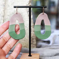Pair of artful taupe and green earrings made by Jax Atelier on a black stand held by a hand.