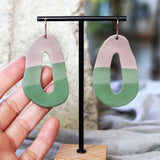 Pair of artful taupe and green earrings made by Jax Atelier on a black stand held by a hand.