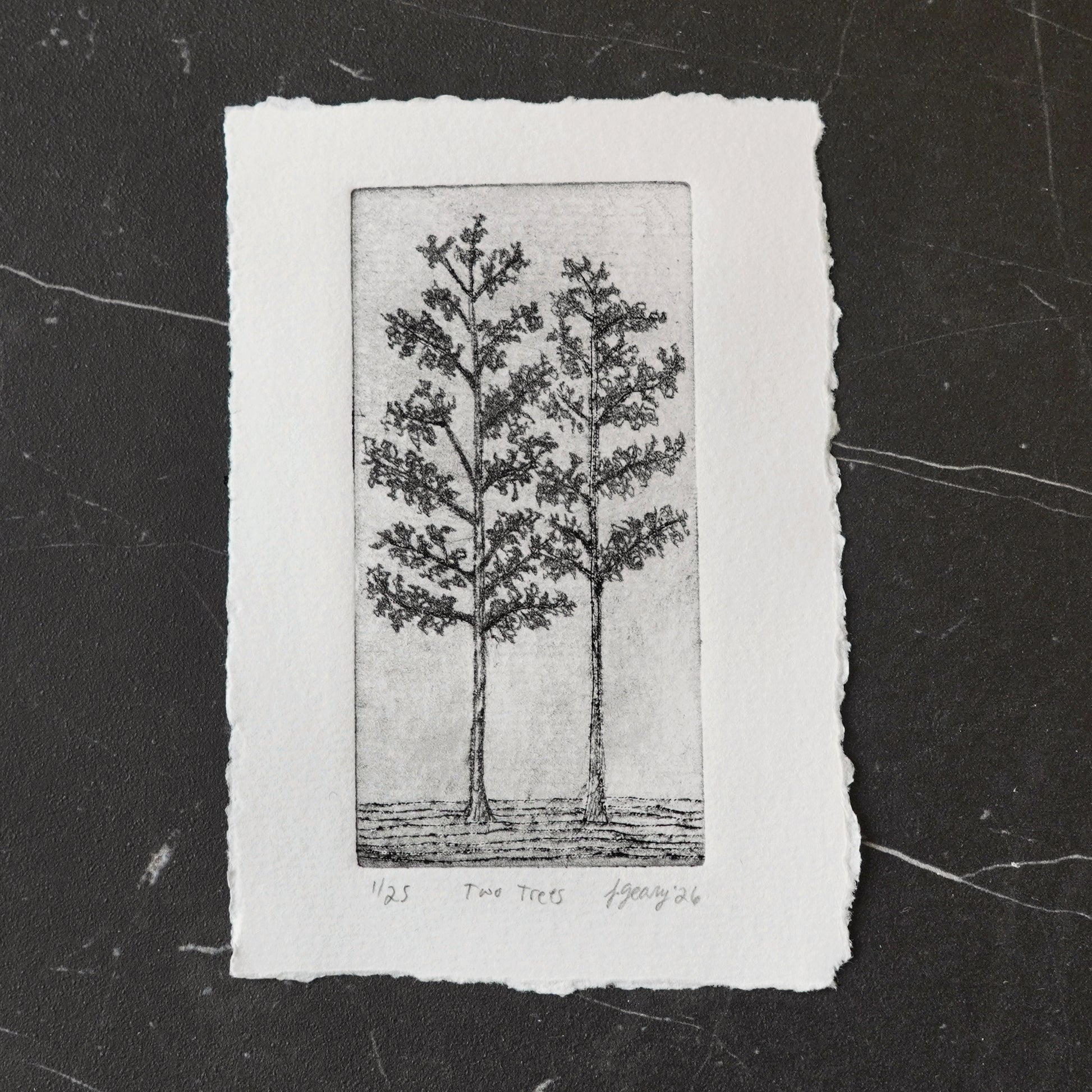 A black and white drypoint etching of two trees on deckled edge white watercolor paper, displayed on a dark marbled tile surface. By Jax Atelier in San DIego.